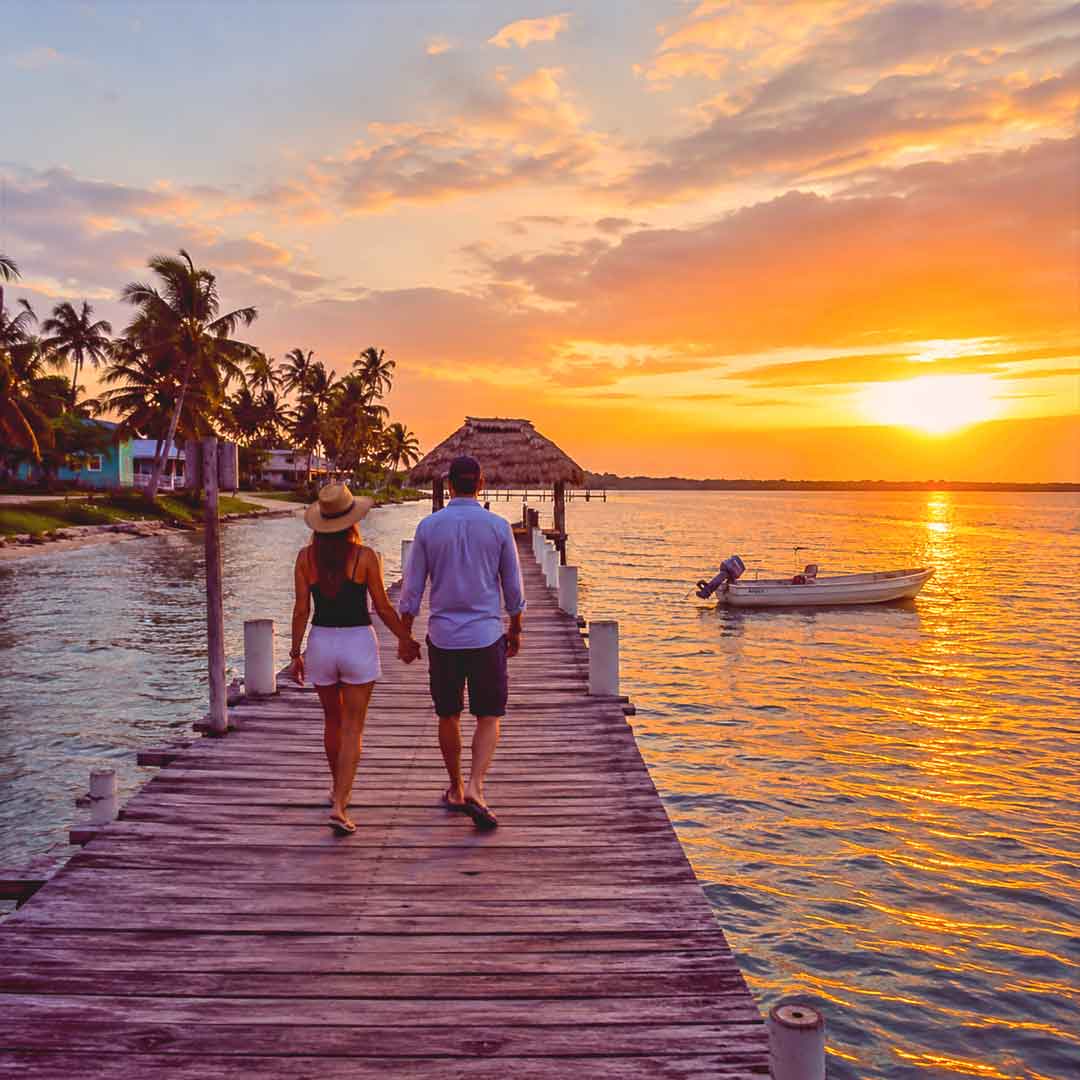 Belize-Destinations-Edit - Authentic Travel Belize Couple walking hand in hand along a wooden pier at sunset in Belize surrounded by turquoise Caribbean waters, tropical palms, and a peaceful island atmosphere.