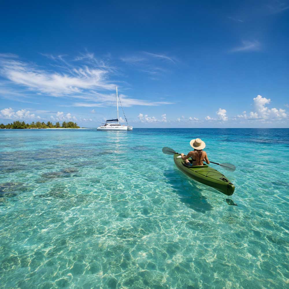 Kaking-The-Reef - Authentic Travel Belize Woman kayaking in turquoise Caribbean waters during a Belize sailing vacation with a luxury catamaran anchored in the distance near the Belize Barrier Reef.