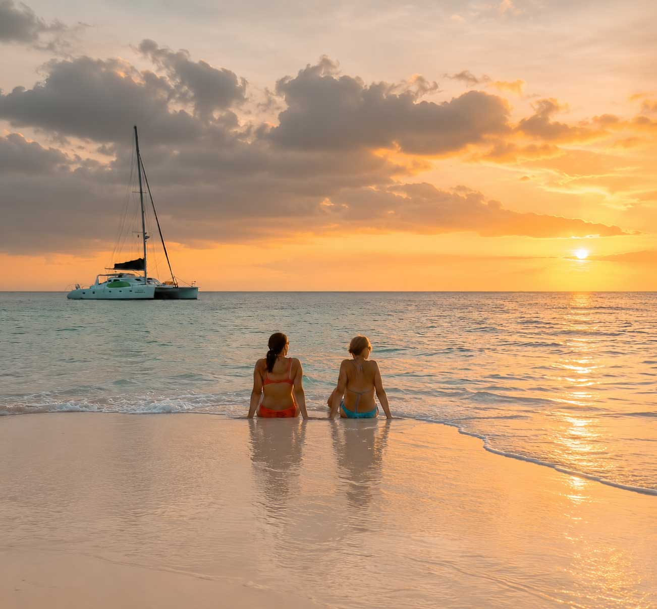 Sunset-Saliling-Belize-Vacations - Authentic Travel Belize Two women enjoying a peaceful sunset beach moment during Belize Sailing Vacations with a luxury catamaran anchored offshore in the Caribbean Sea.