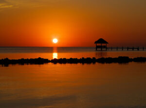 Sunset-Corozal-Bay- - Authentic Travel Belize Sunset over calm water with an orange sky and a thatched gazebo on a pier to the right, silhouetted against the horizon.