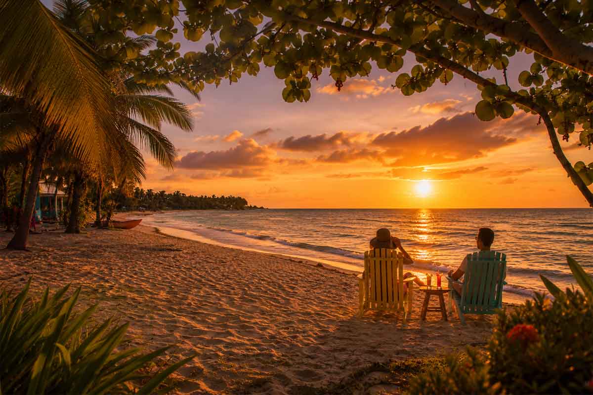 Belize-honeymoon-and-romantic-scapes - Authentic Travel Belize Couple seated on a Belize beach enjoying tropical drinks at Caribbean sunset, showcasing romantic Belize travel styles for a relaxed coastal escape.