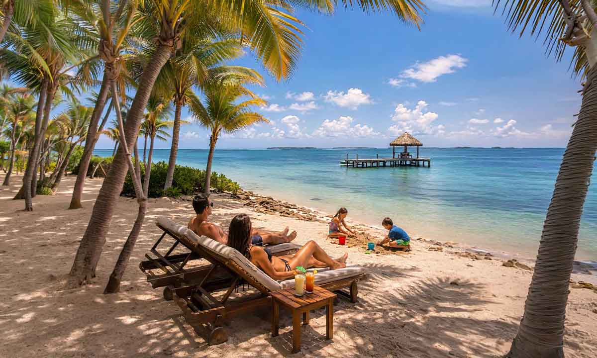 Coast-&-Relaxation-Family-Journey - Authentic Travel Belize Belize Family Vacations with parents relaxing in beach chairs while children play in the sand beside clear turquoise water and the Belize Barrier Reef.