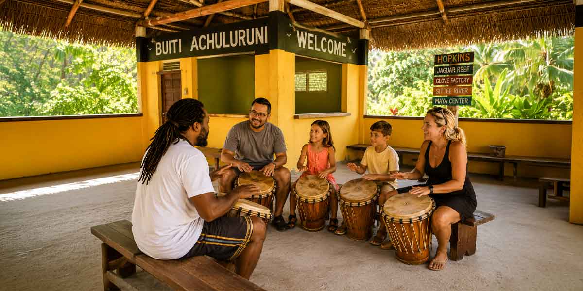 Cultural-Adventure-Belize - Authentic Travel Belize Group of people (adults and children) sitting on benches in a yellow open-air shelter, playing djembes together in a drumming circle.