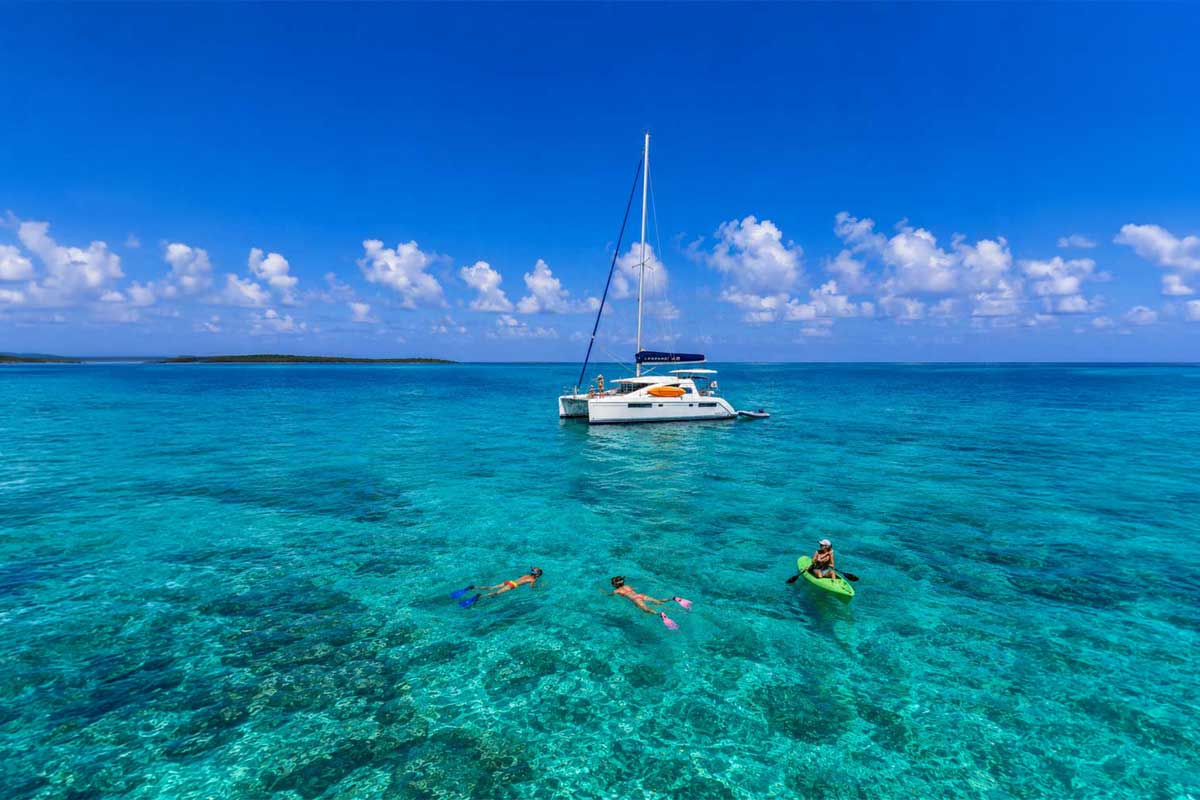 Honemoon-Placencia - Authentic Travel Belize Mother kayaking beside her child snorkeling in turquoise Belize waters, with a catamaran in the background, highlighting family-friendly Belize travel styles.